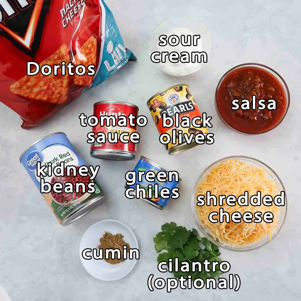 Overhead shot of ingredients - doritos, sour cream, kidney beans, tomato sauce, black olives, salsa, green chiles, shredded cheese, cilantro, and cumin.