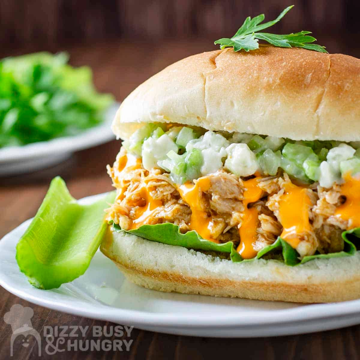 Side shot of buffalo chicken sandwiches garnished with fresh herbs with a celery stick on the side on a white plate.