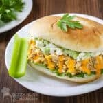 Side angled shot of buffalo chicken sandwiches garnished with fresh herbs with a celery stick on the side on a white plate.
