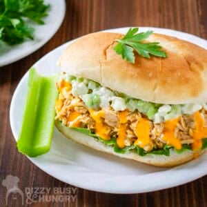 Side angled shot of buffalo chicken sandwiches garnished with fresh herbs with a celery stick on the side on a white plate.