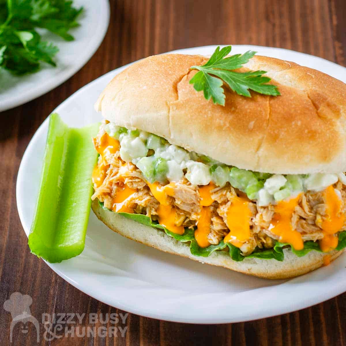 Side angled shot of buffalo chicken sandwiches garnished with fresh herbs with a celery stick on the side on a white plate.