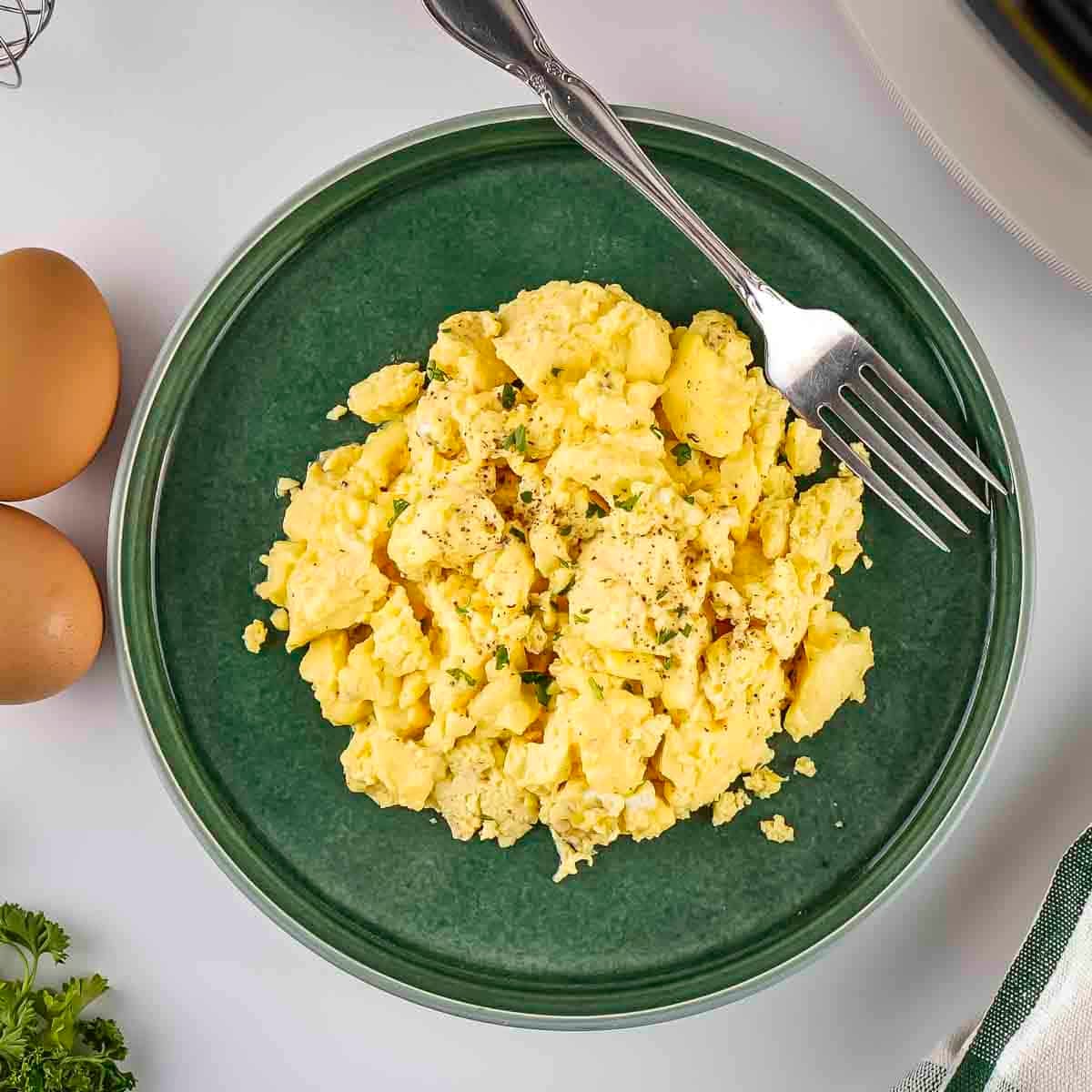 Overhead shot of scrambled eggs garnished with fresh herbs on a sage green plate with a fork on the side and eggs in the background.