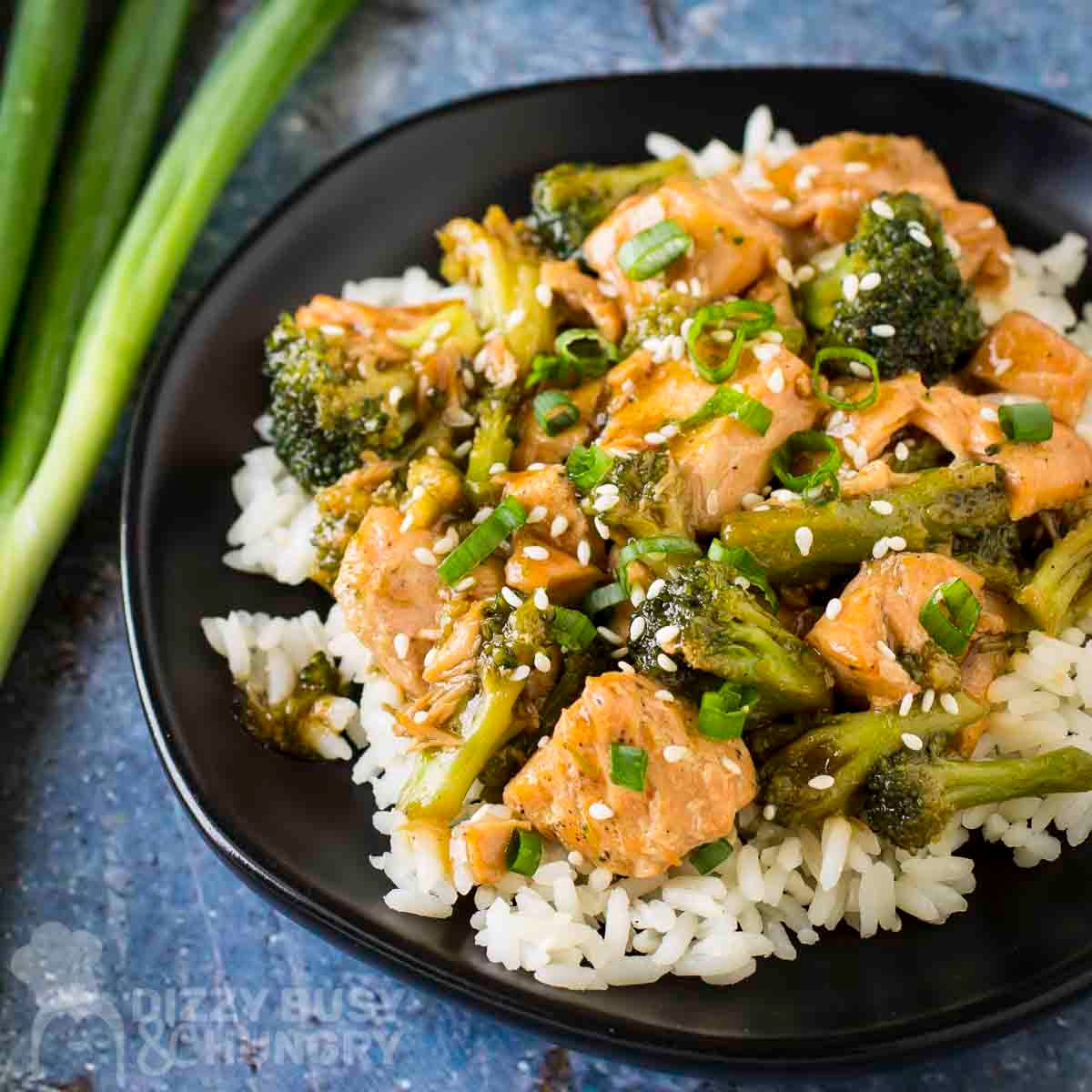 Side shot of teriyaki salmon and broccoli on a bed or rice garnished with fresh green onions on a black plate with more green onions on the side.