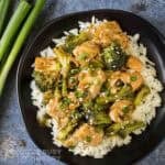 Overhead shot of teriyaki salmon and broccoli on a bed or rice garnished with fresh green onions on a black plate with more green onions on the side.