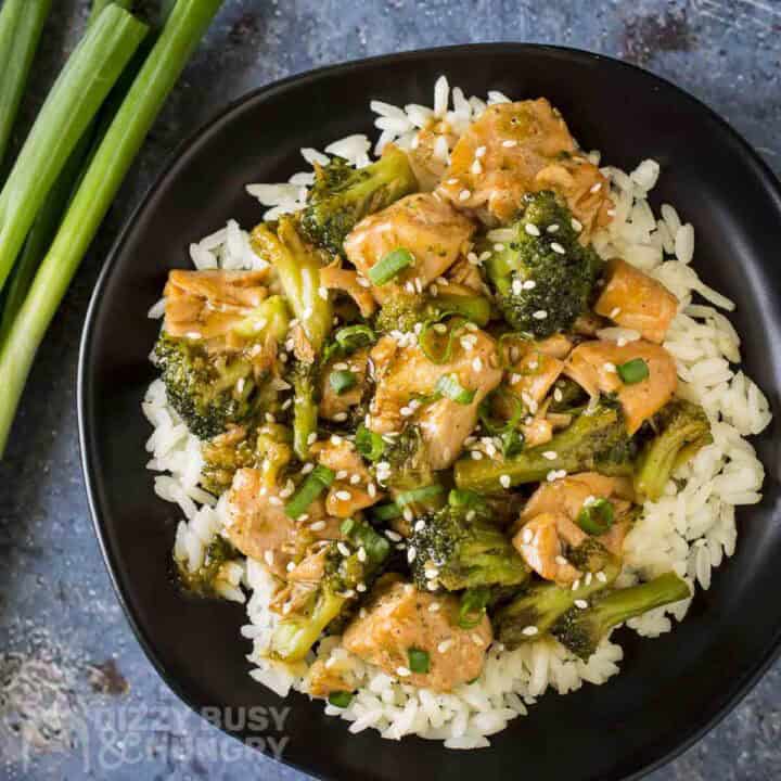 Overhead shot of teriyaki salmon and broccoli on a bed or rice garnished with fresh green onions on a black plate with more green onions on the side.