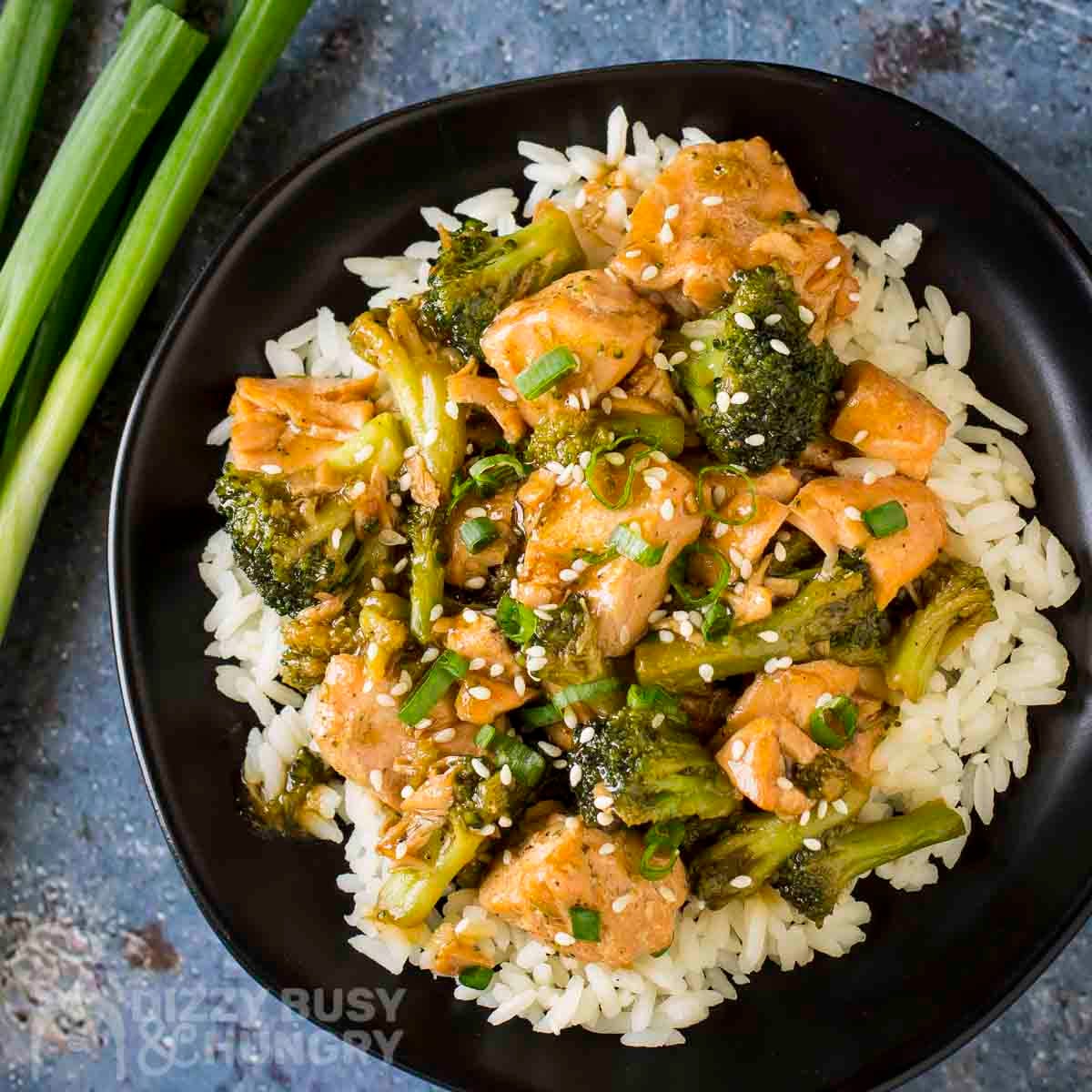 Overhead shot of teriyaki salmon and broccoli on a bed or rice garnished with fresh green onions on a black plate with more green onions on the side.