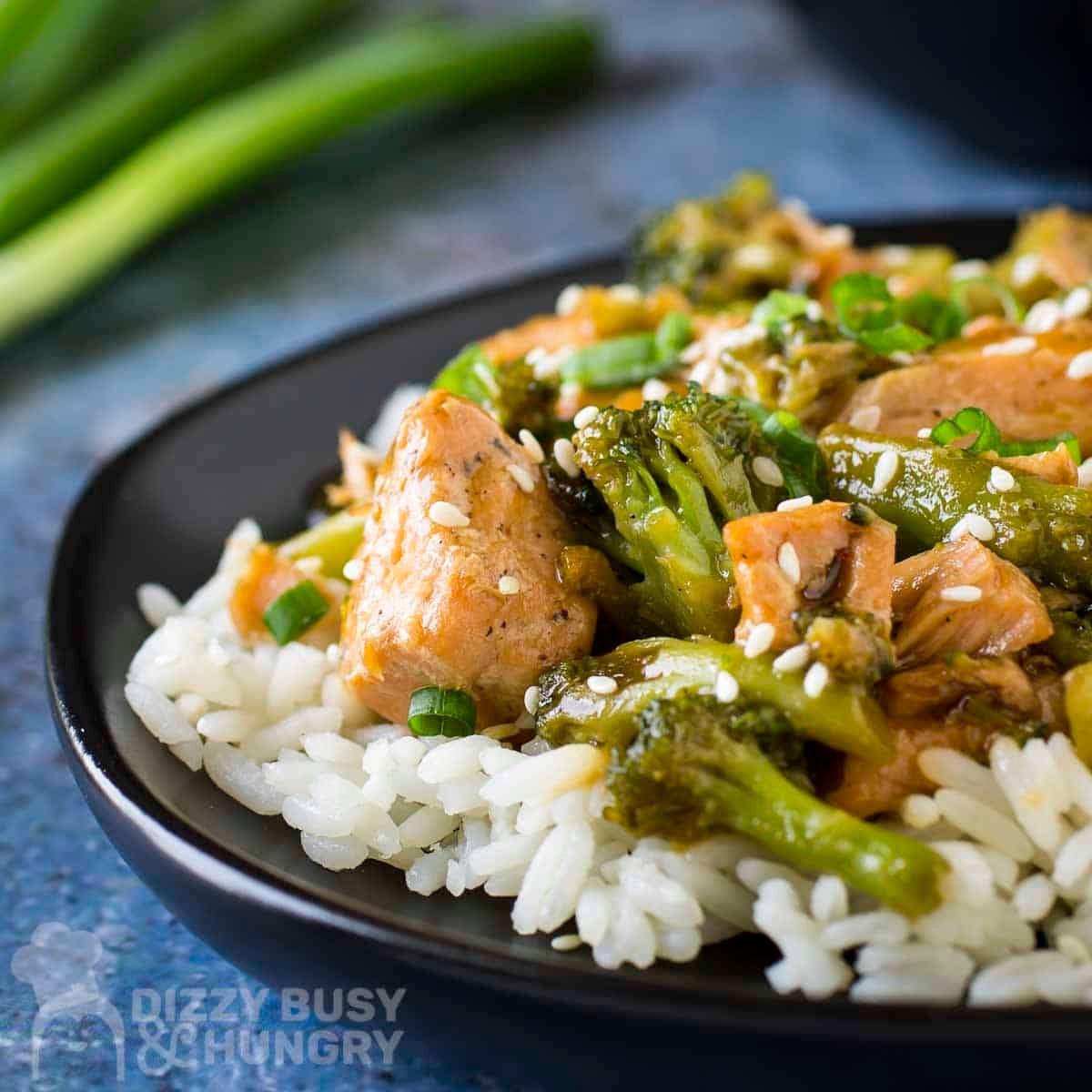 Side close up shot of teriyaki salmon and broccoli on a bed or rice garnished with fresh green onions on a black plate with more green onions on the side.