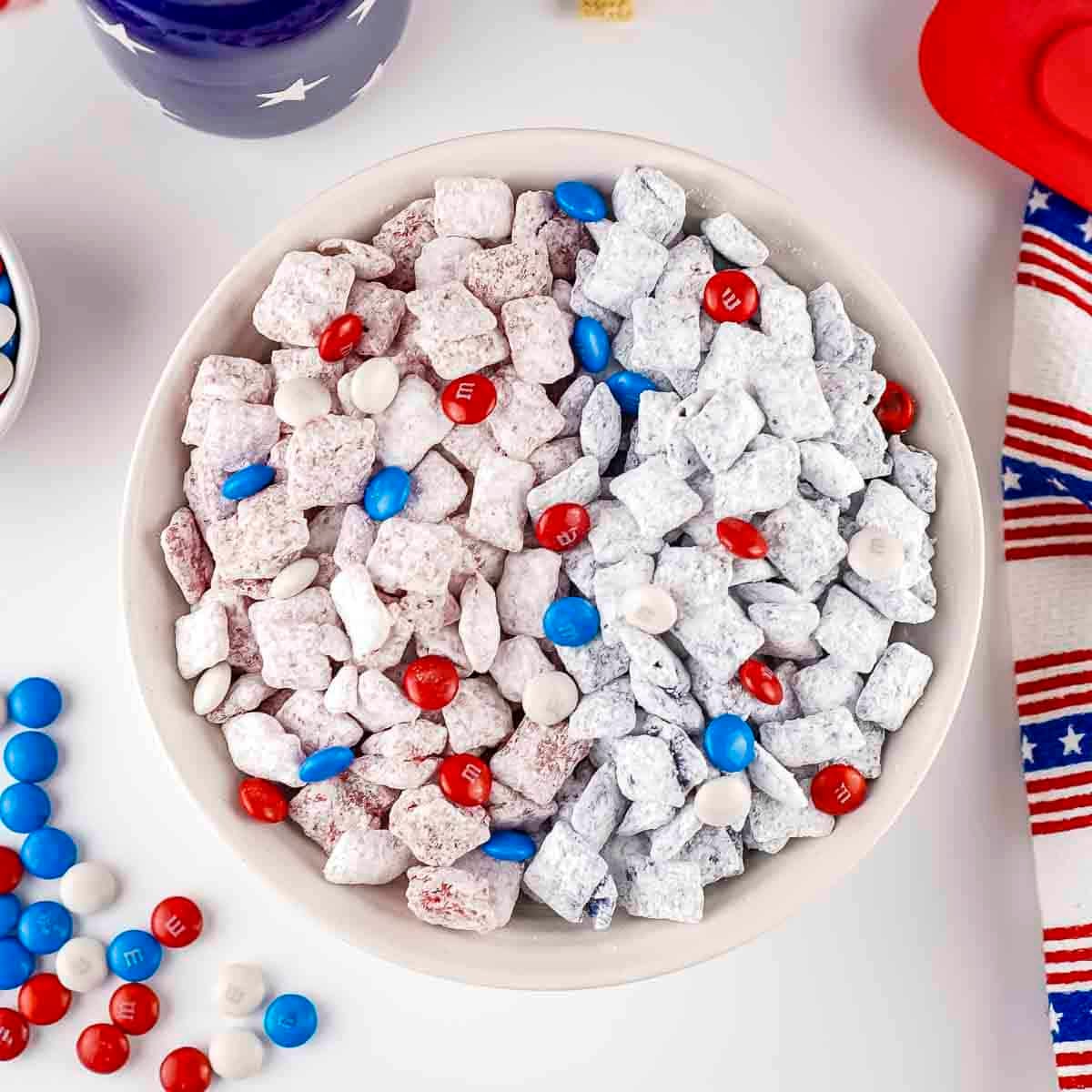 Overhead shot of patriotic muddy buddies in a white bowl on a white surface with blue and white bowls and napkins in the background.