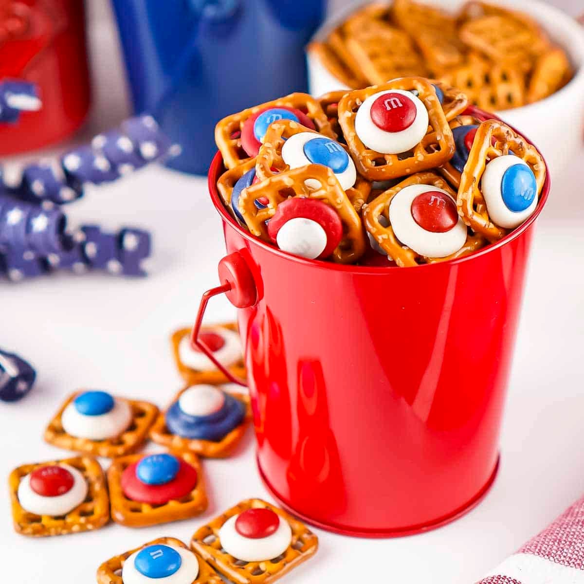 Side shot of patriotic pretzels in a red bucket with blue ribbon and red and blue buckets in the background.