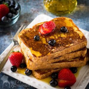 Side shot of french toast stacked on a white plate garnished with sliced strawberries, blueberries, and syrup with a fork on the side.