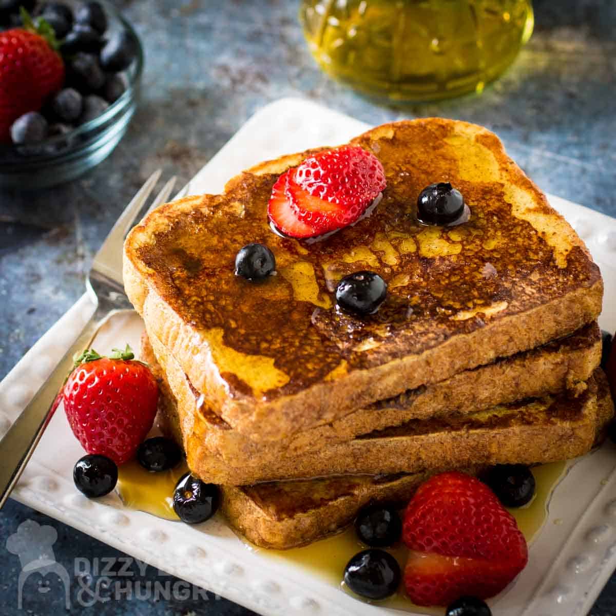 Side shot of french toast stacked on a white plate garnished with sliced strawberries, blueberries, and syrup with a fork on the side.