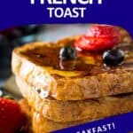 Side shot of french toast stacked on a white plate garnished with sliced strawberries, blueberries, and syrup.