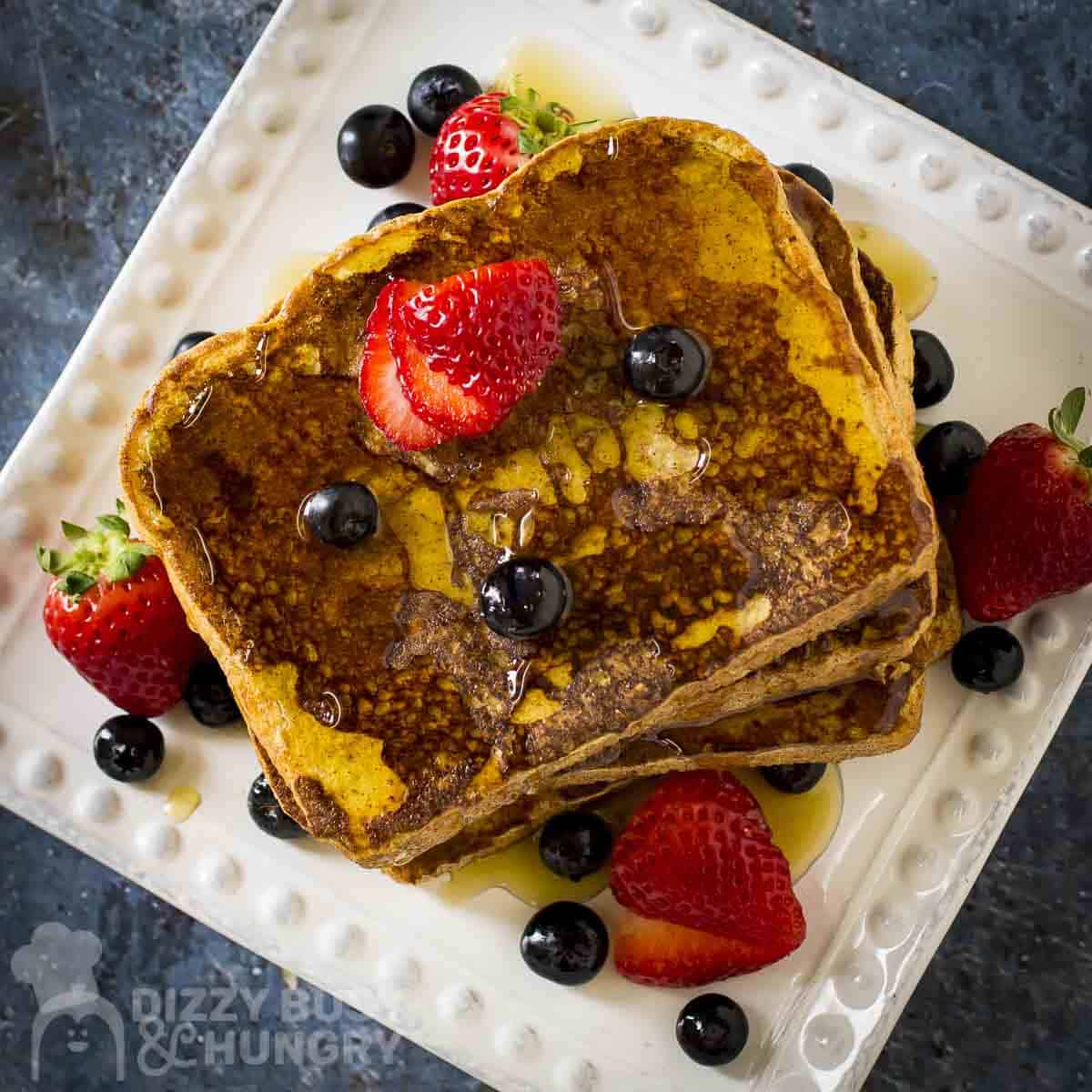 Overhead shot of french toast stacked on a white square plate garnished with sliced strawberries, blueberries, and syrup.