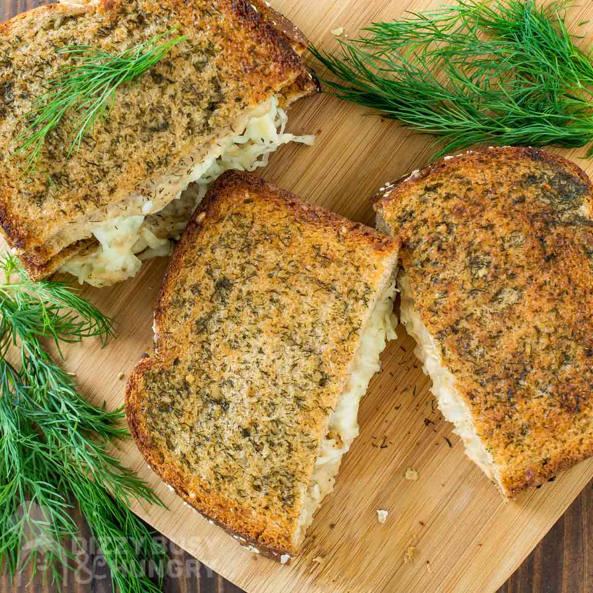 Overhead shot of four grilled cheese halves stacked on a cutting board garnished with fresh dill.