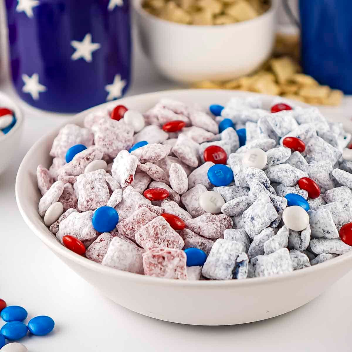 Side shot of patriotic muddy buddies in a white bowl on a white surface with blue and white bowls in the background.
