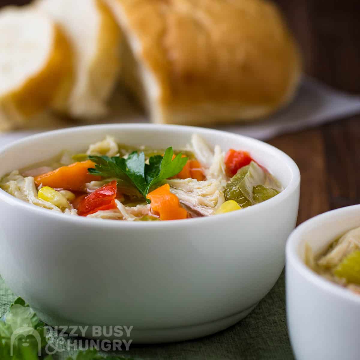Side shot of chicken soup with a sprig of fresh parsley in a white bowl on a green cloth with sliced bread in the background.