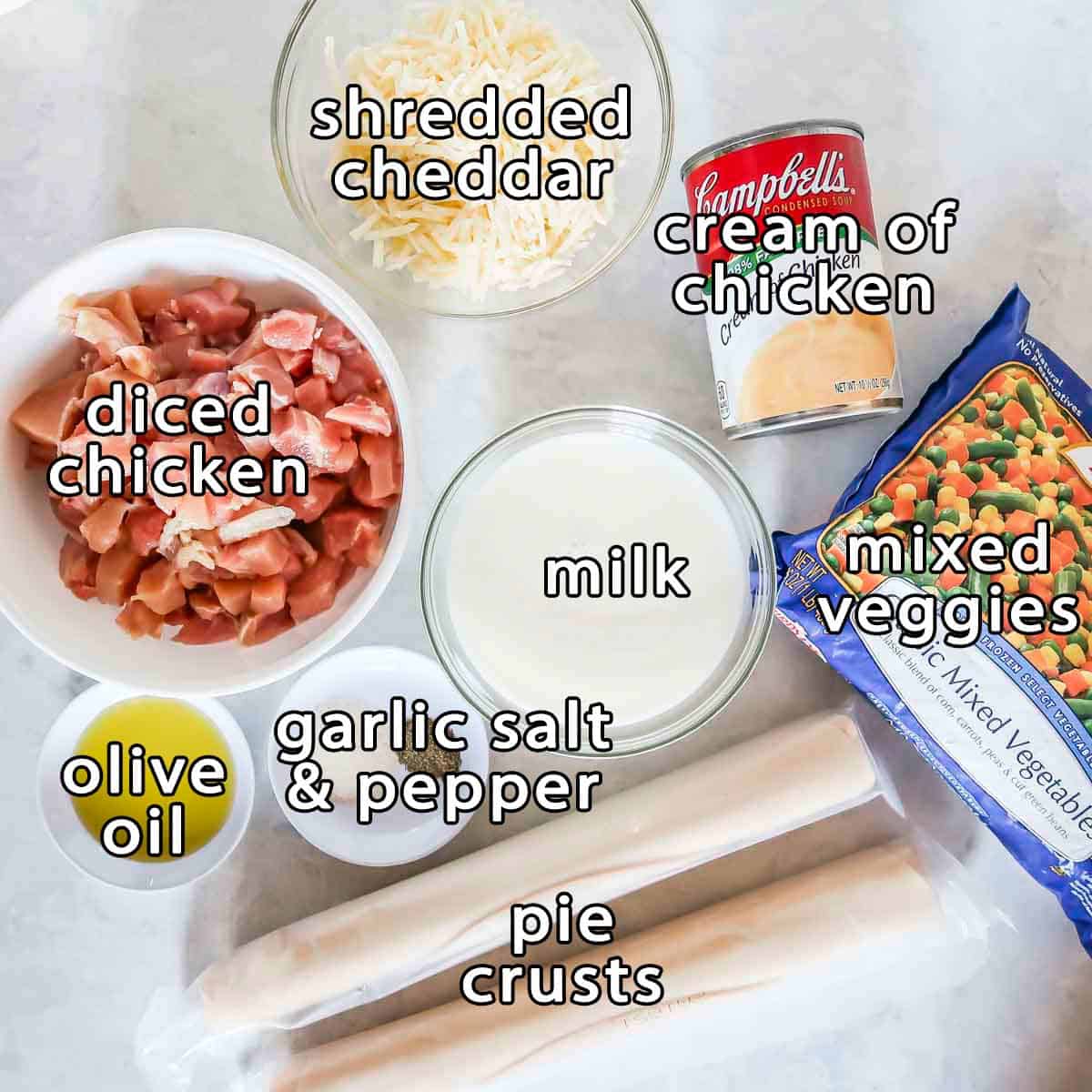 Overhead shot of ingredients - shredded cheddar, diced chicken, cream of chicken, mixed veggies, milk, pie crusts, olive oil, garlic salt, and pepper.