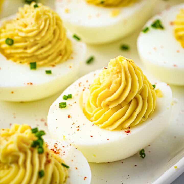 Side close up shot of multiple deviled eggs garnished with paprika and chives on a white plate.