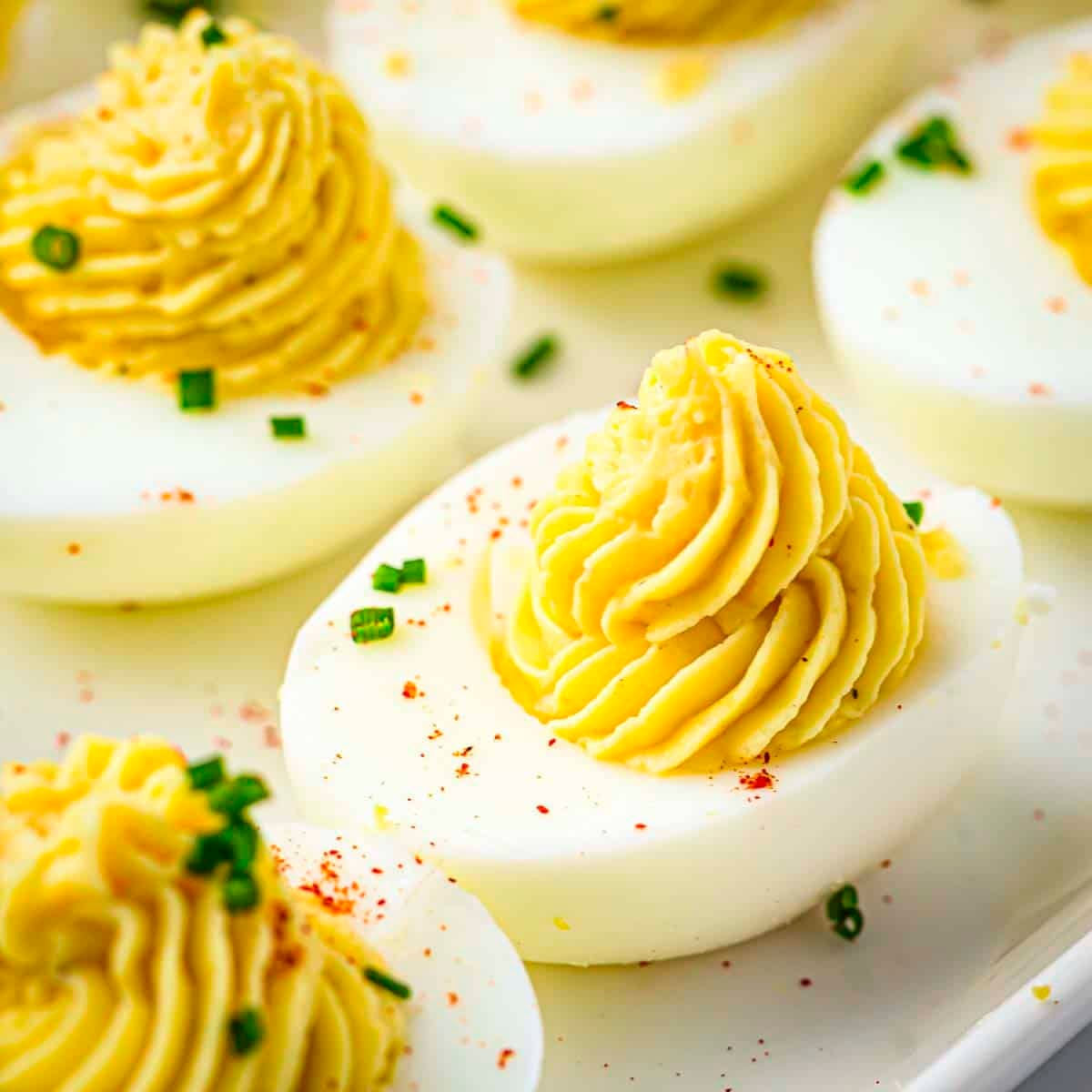 Side close up shot of multiple deviled eggs garnished with paprika and chives on a white plate.
