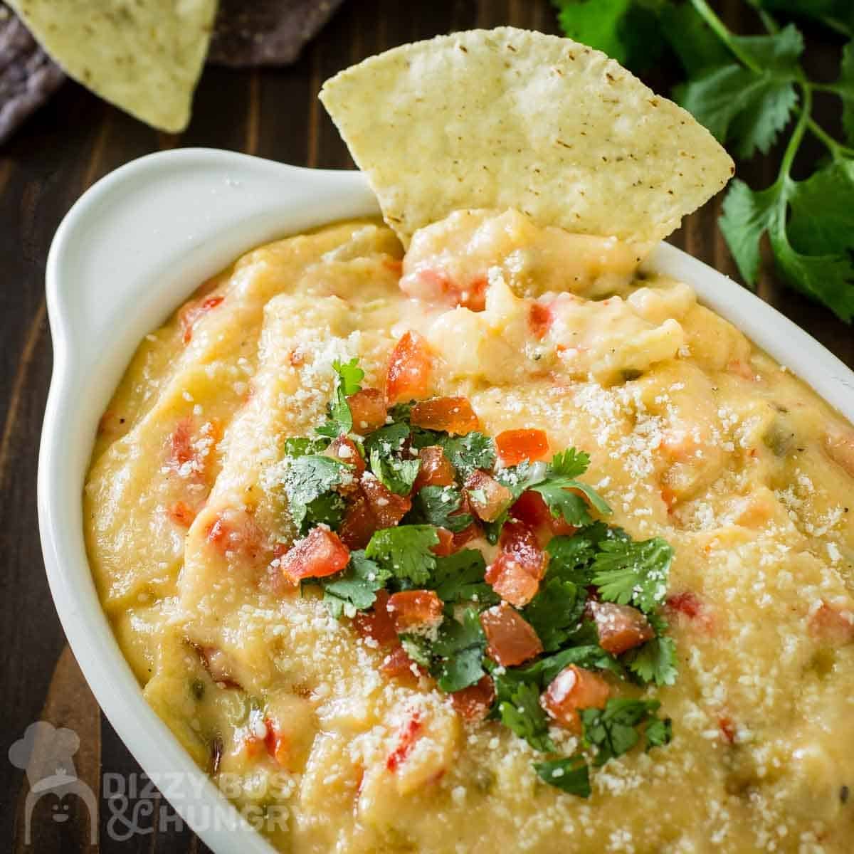 Close up shot of queso dip garnished with parsley and diced tomatoes with a tortilla chip being dipped in.