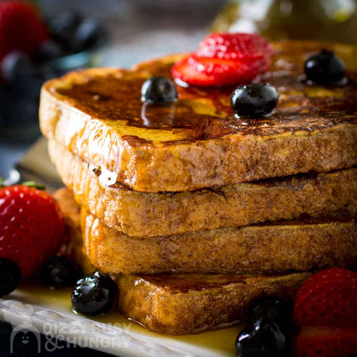 Side close up shot of french toast stacked on a white plate garnished with sliced strawberries, blueberries, and syrup with a fork on the side.