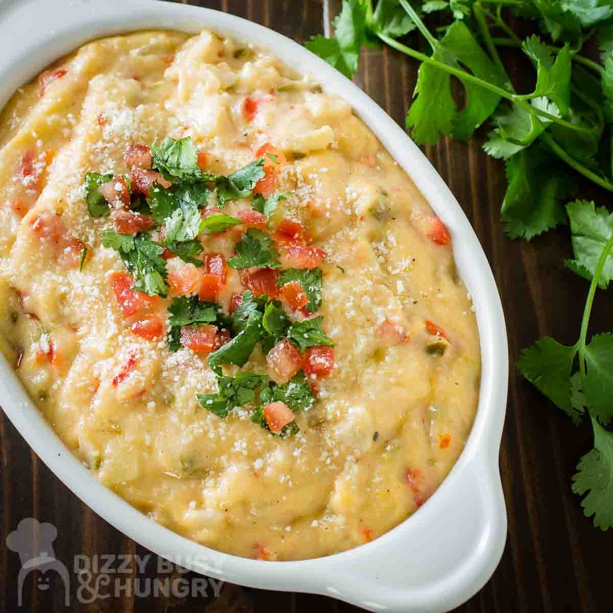 Close up shot of queso dip in a white serving bowl garnished with parsley and diced tomatoes with a bundle of fresh parlsey on the side.