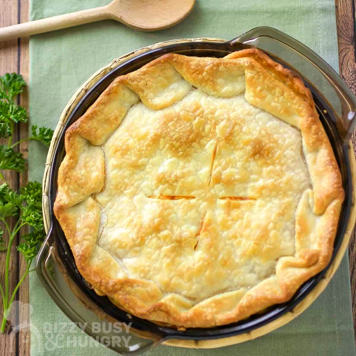 Overhead view of golden brown chicken pot pie in a casserole dish on a green cloth with a wooden spoon and fresh parsley on the side.