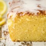 Side shot of lemon loaf cake sliced in the middle with lemon glaze on a wooden surface with sliced lemons on the side.