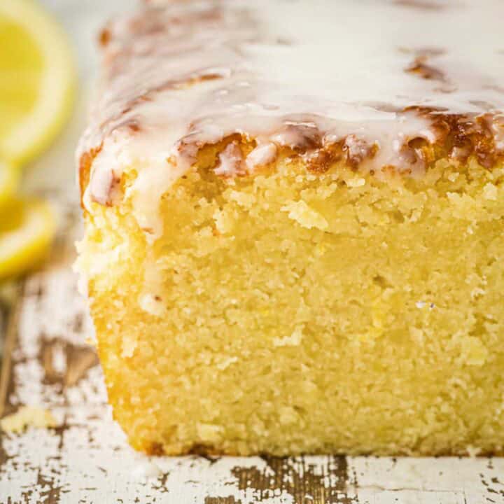 Side shot of lemon loaf cake sliced in the middle with lemon glaze on a wooden surface with sliced lemons on the side.