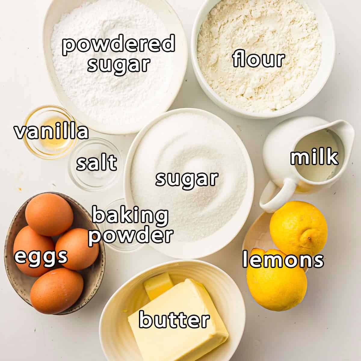 Overhead view of ingredients - powdered sugar, flour, sugar, eggs, baking powder, salt, vanilla, milk, lemons, and butter.