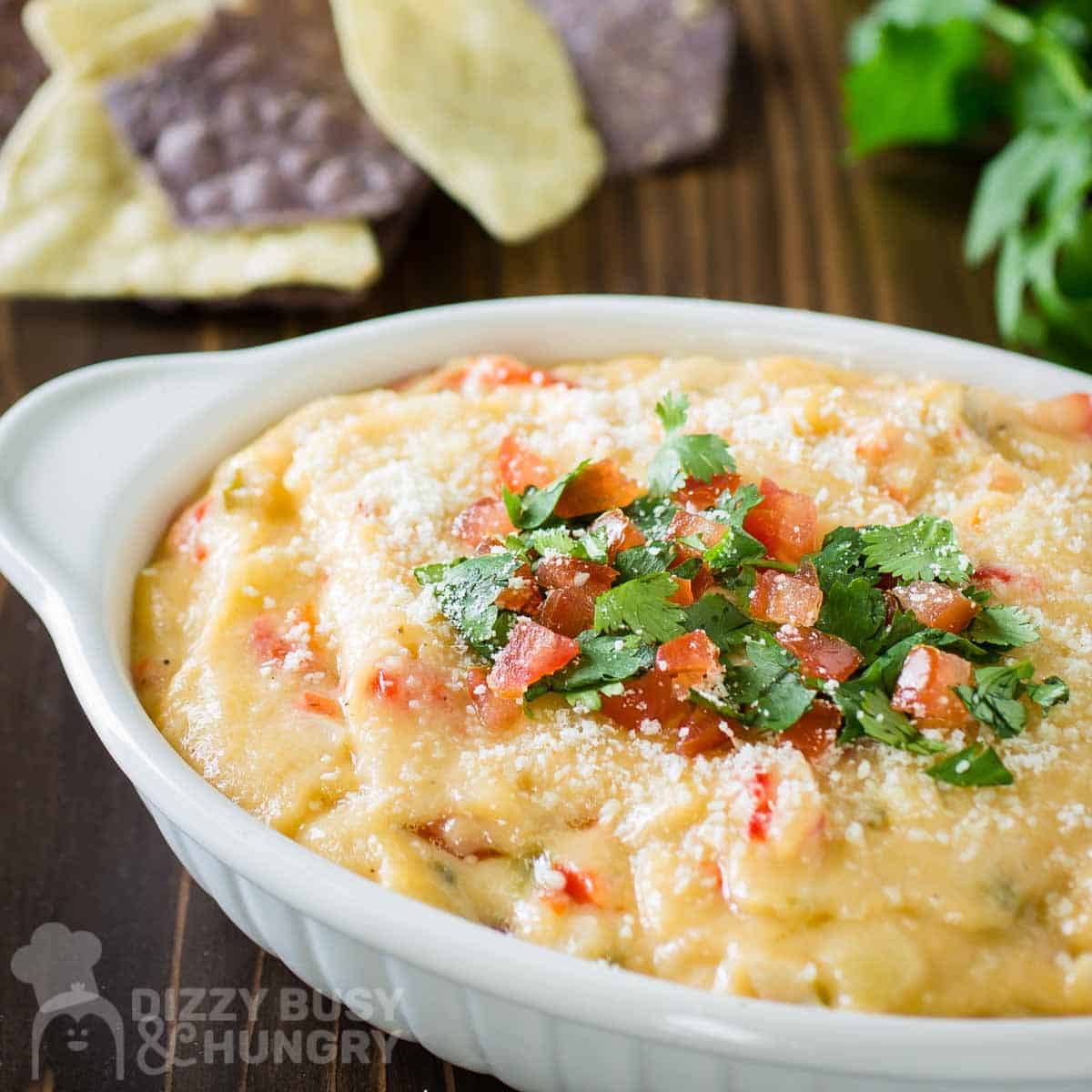 Close up shot of queso dip garnished with parsley and diced tomatoes with fresh parsley and tortilla chips in the background.