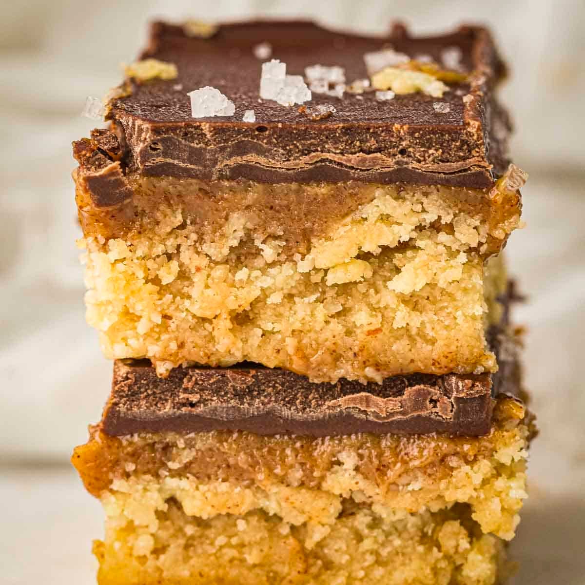 Side shot of two caramel bars stacked on each other sprinkled with flaky salt on a white surface.