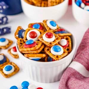 Close up shot of patriotic pretzels in a white ramekin on a white surface with more pretzels and M&Ms in white bowls in the background.