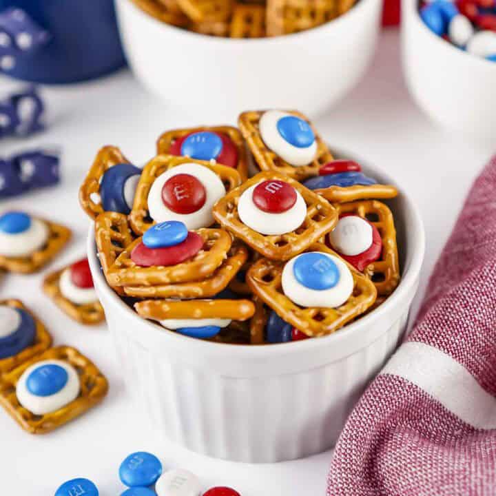 Close up shot of patriotic pretzels in a white ramekin on a white surface with more pretzels and M&Ms in white bowls in the background.