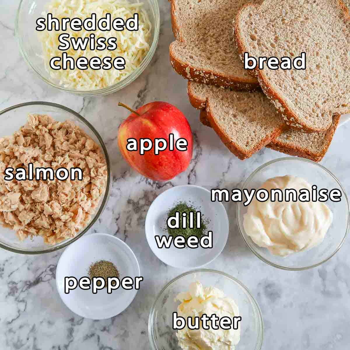 Overhead shot of ingredients - salmon, shredded swiss cheese, bread, apple, dill weed, butter, mayonnaise, and pepper.