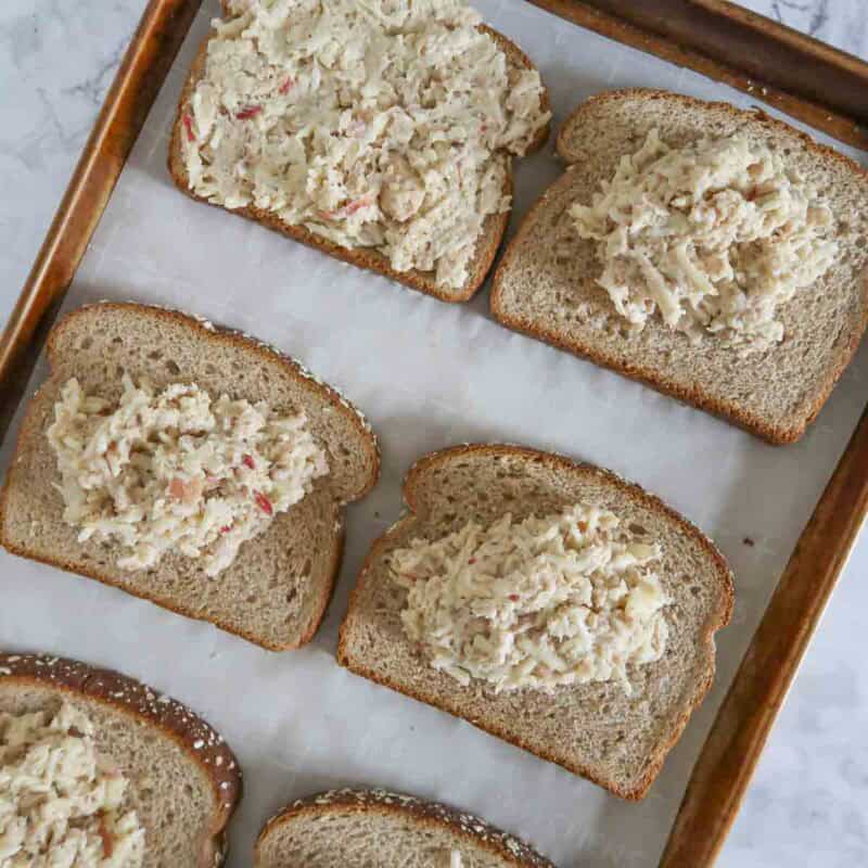 Overhead view of process shot 3 - butter one side of each slice of bread with the dill weed mixture. Place bottom slices buttered side down on the baking sheet. Distribute the salmon mix over the bottom slices and spread it out.