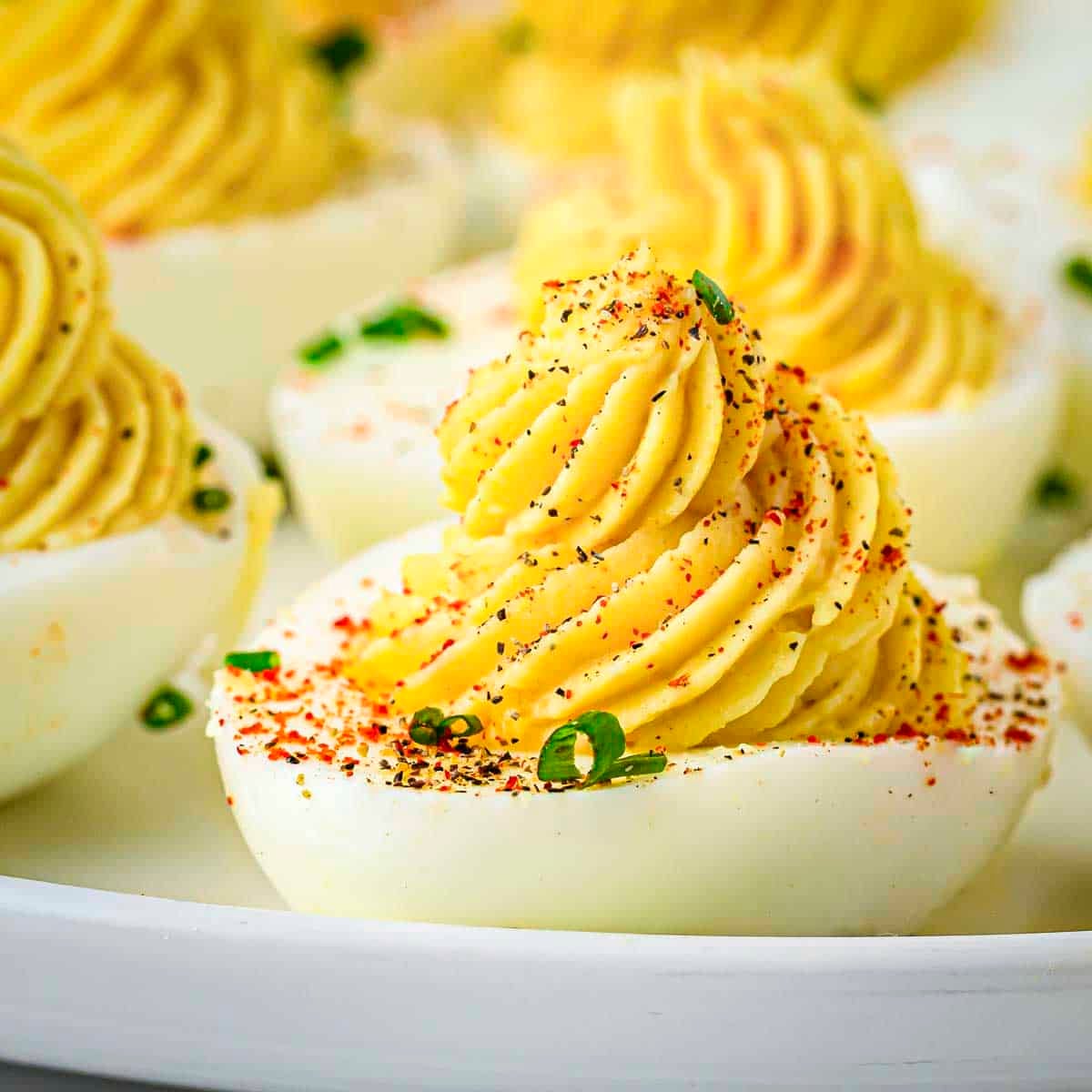 Side shot of multiple deviled eggs garnished with paprika and chives on a white plate.