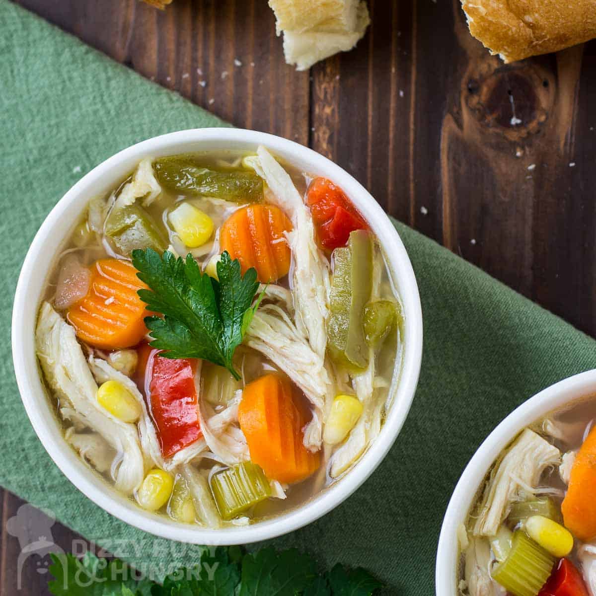 Overhead shot of chicken soup with a sprig of fresh parsley in a white bowl on a green cloth with pieces of bread on the side.
