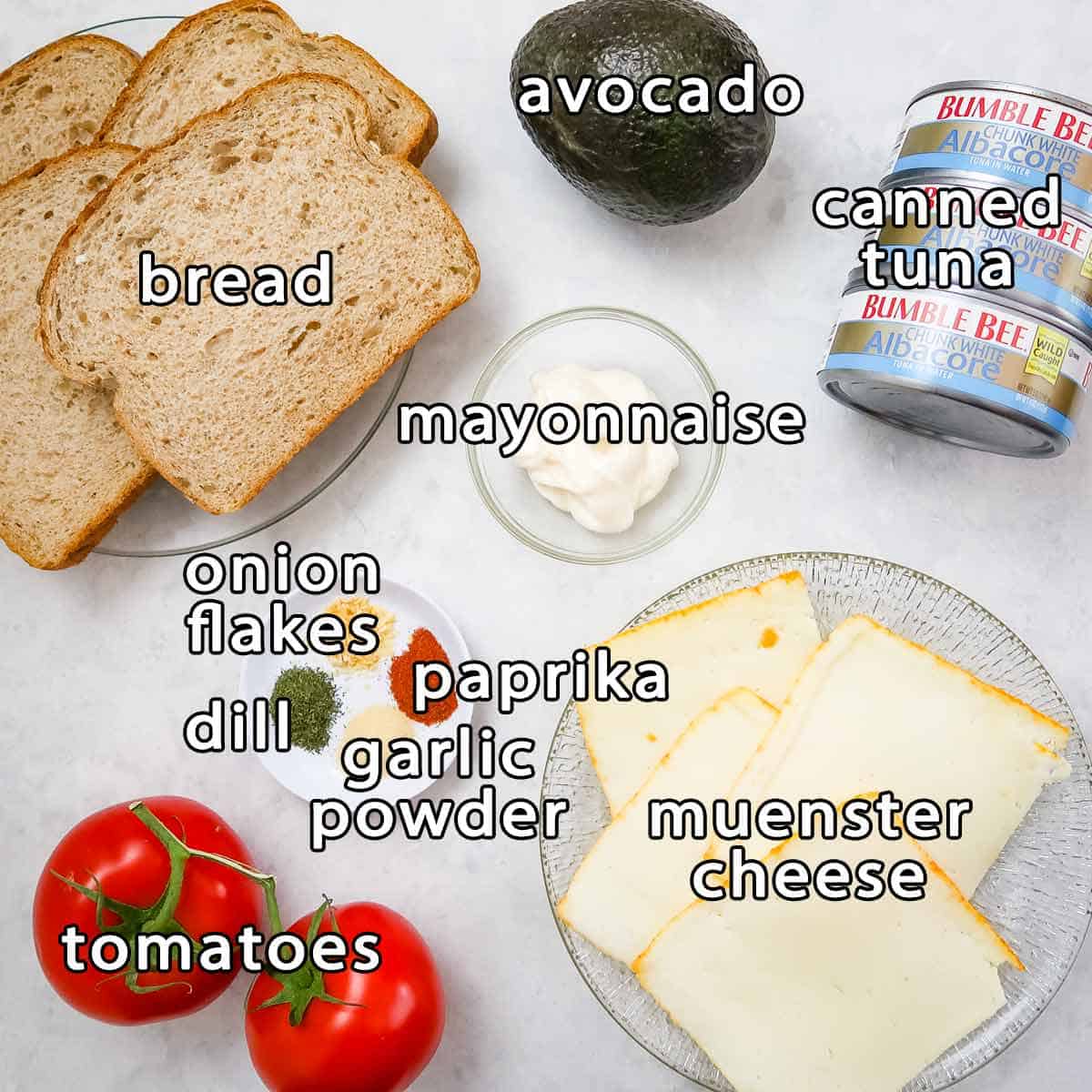 Overhead view of ingredients - bread, avocado, canned tuna, mayonnaise, muenster cheese, onion flakes, paprika, garlic powder, dill, and tomatoes.
