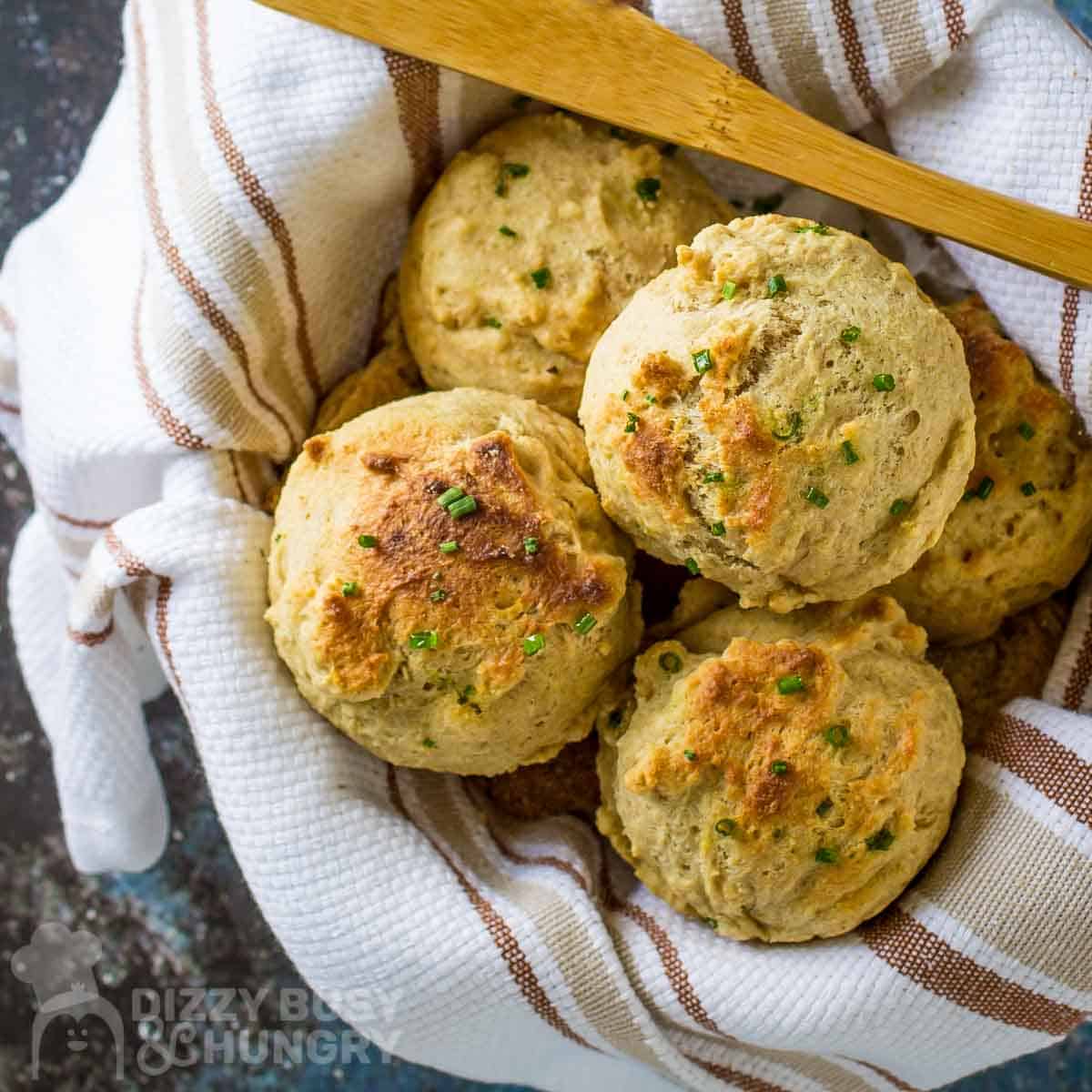 Overhead shot of golden brown biscuits garnished with chives in a bowl wrapped in a white striped cloth with a wooden spoon on the side.