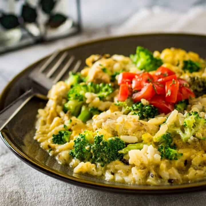 Side shot of cheesy chicken and rice garnished with herbs and fresh tomatoes in a brown bowl with a fork on the side.