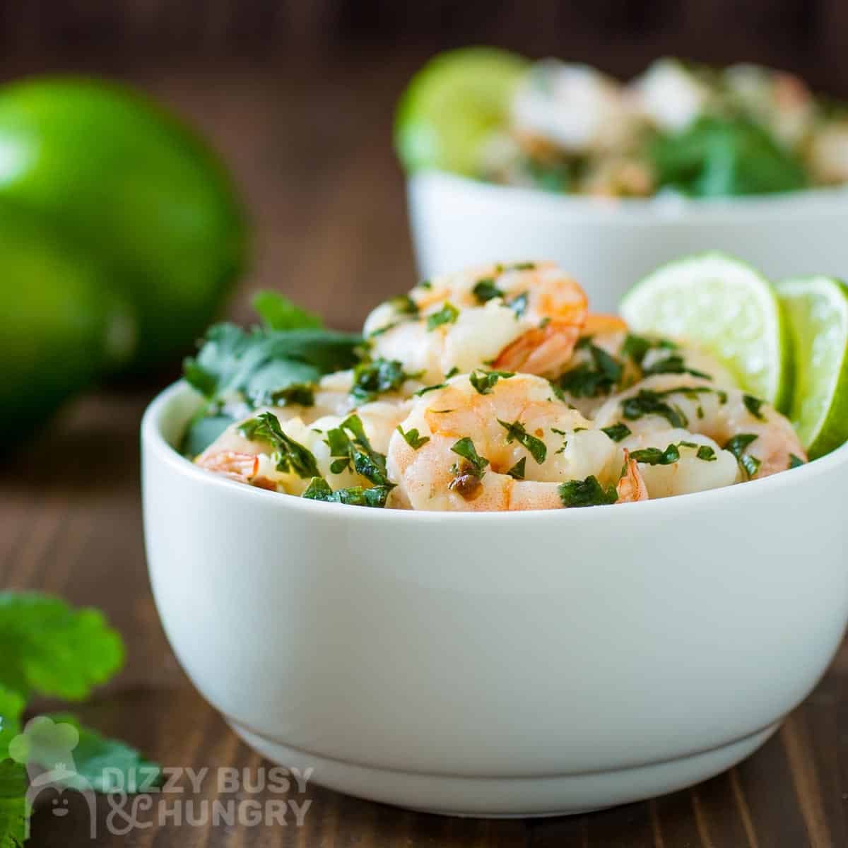 Side shot of butter lime shrimp garnished with fresh parsley and lime wedges in a white bowl with more limes and parsley sprigs on the side.