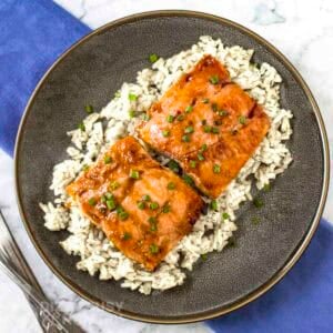 Overhead shot of glazed salmon on a bed of rice garnished with fresh green onions in a black bowl.
