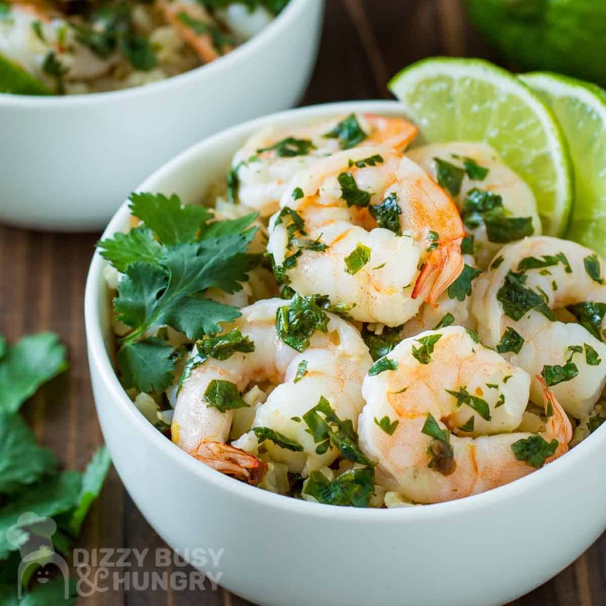 Close up shot of butter lime shrimp garnished with fresh parsley and lime wedges in a white bowl with more limes and parsley sprigs on the side.