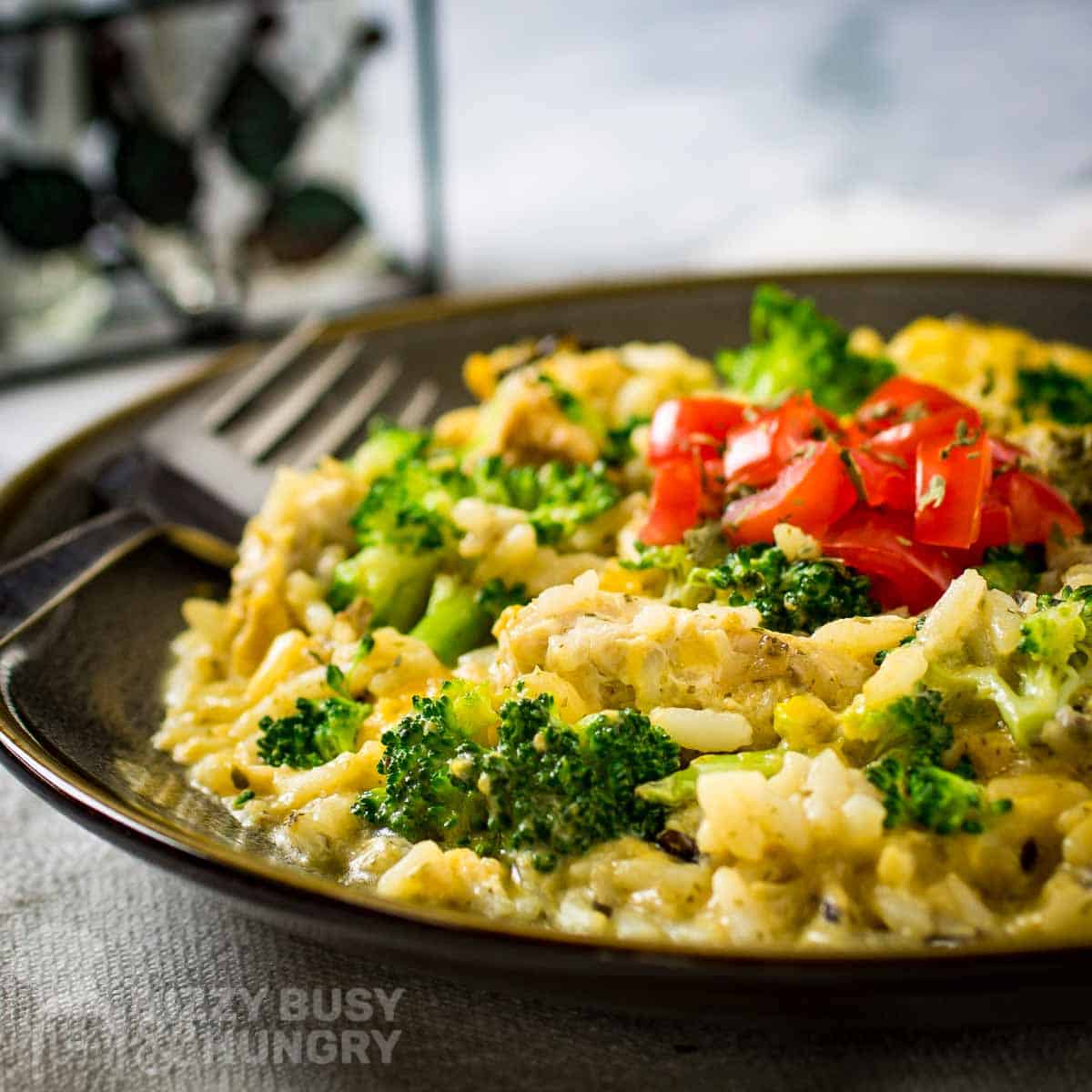 Side shot of cheesy chicken and rice garnished with herbs and fresh tomatoes in a brown bowl with a fork on the side.