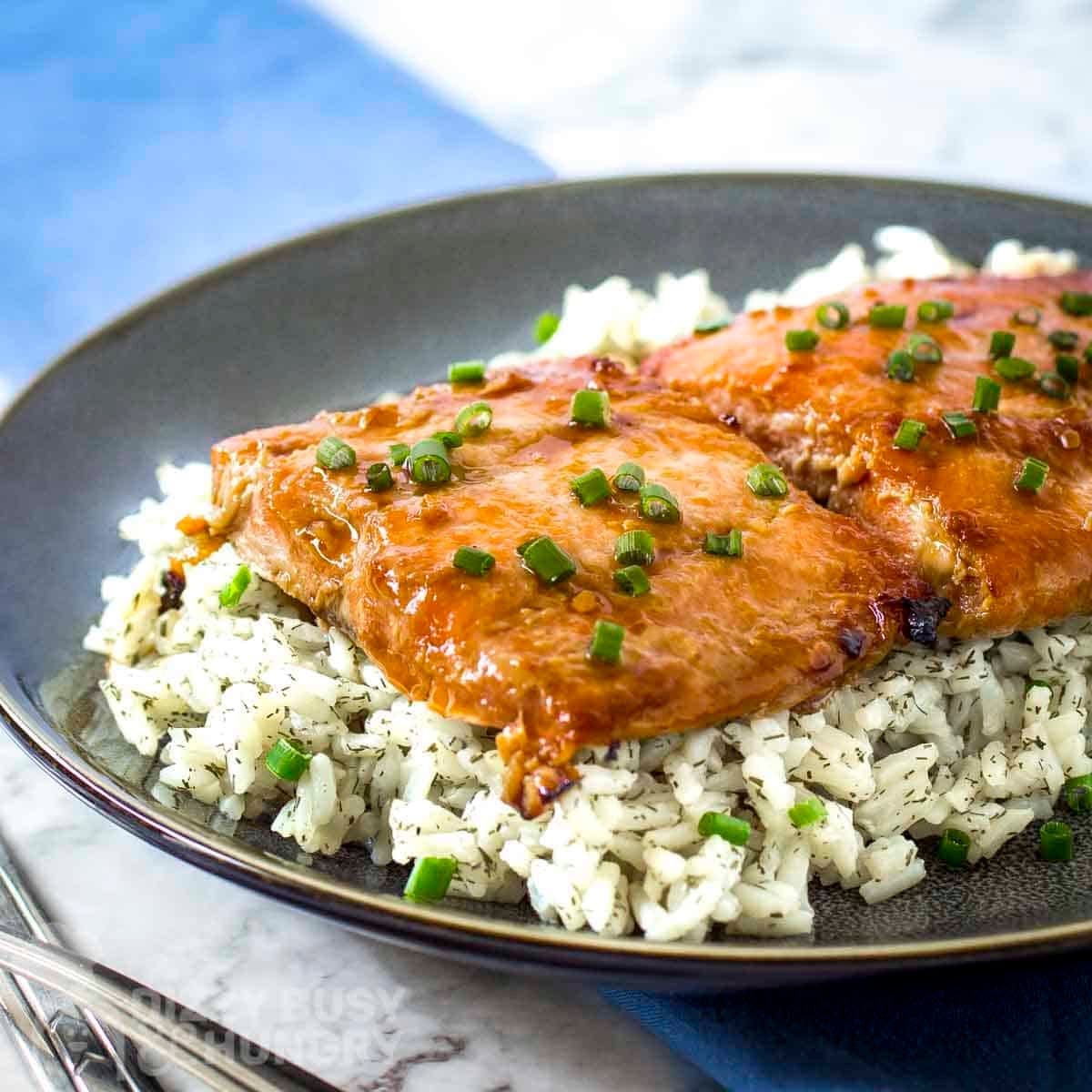 Side shot of glazed salmon on a bed of rice garnished with fresh green onions in a black bowl.