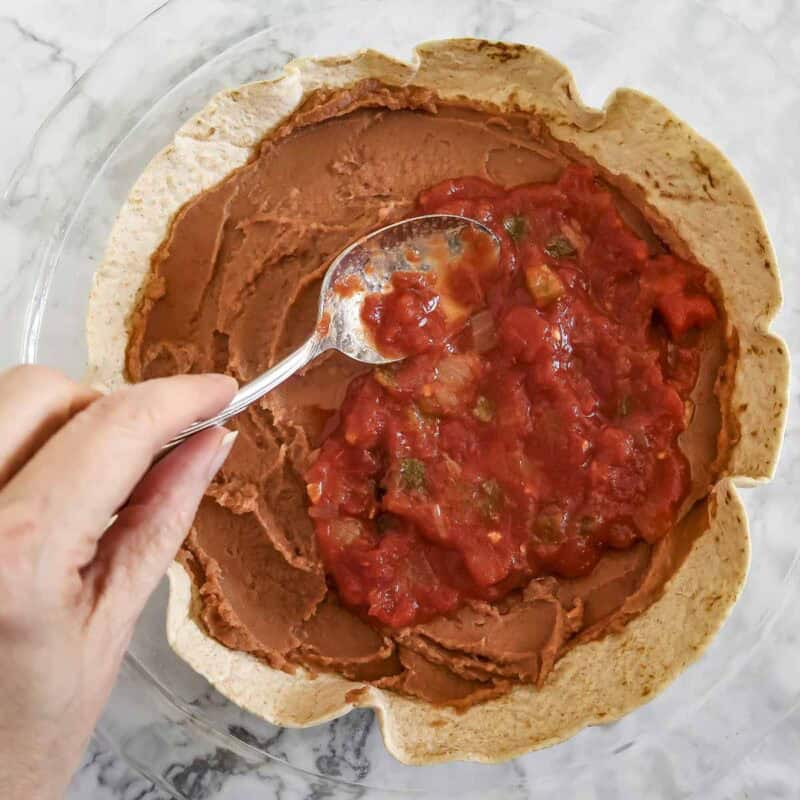 Overhead view of process shot 1 - place one of the tortillas in the bottom of the pan, letting the edges go up the sides. Spread about half of the refried beans on the tortilla, then about a third of the salsa.