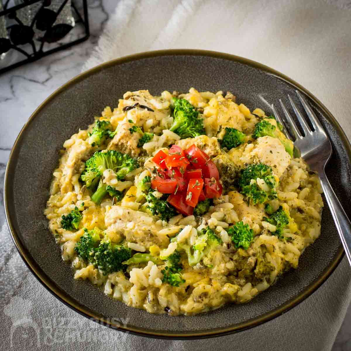 Side angled shot of cheesy chicken and rice garnished with herbs and fresh tomatoes in a brown bowl with a fork on the side.