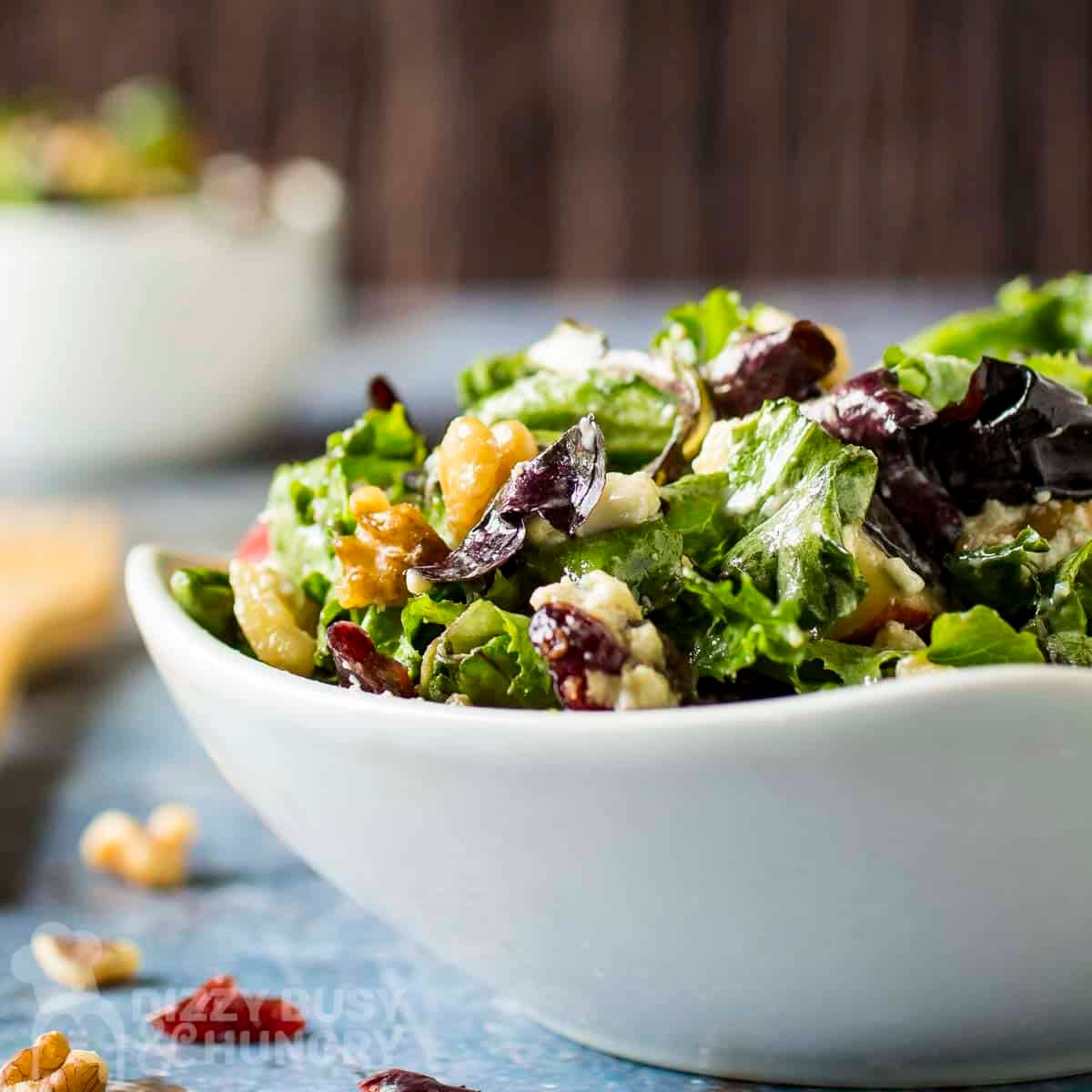 Side shot of apple walnut salad in a white bowl with cranberries, walnuts, and wooden cutlery on the side.