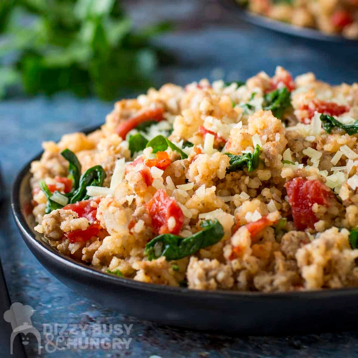 Side shot of couscous and ground turkey on a black plate with fresh herbs in the background.